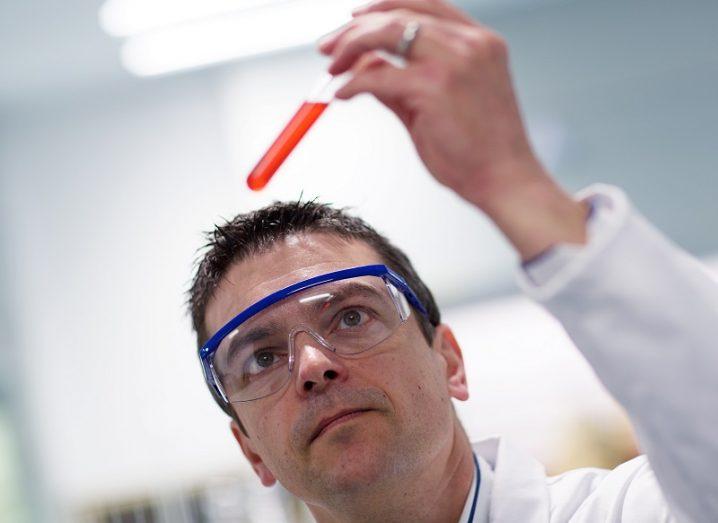 Marc Devocelle wearing safety glasses while holding up a test tube containing a red liquid.