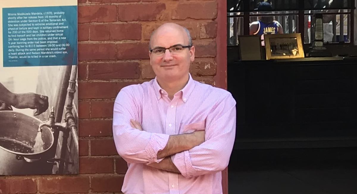 Mike Feerick is wearing a pink shirt while standing in front of a red brick wall and smiling into the camera.