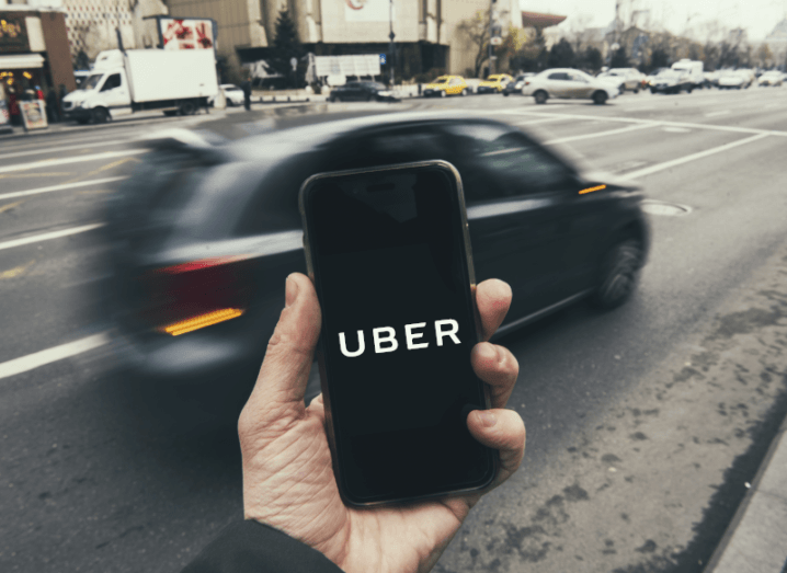 A person holding a smartphone in front of them with the Uber logo displayed on the screen as a car goes by quickly on the street ahead of the phone.