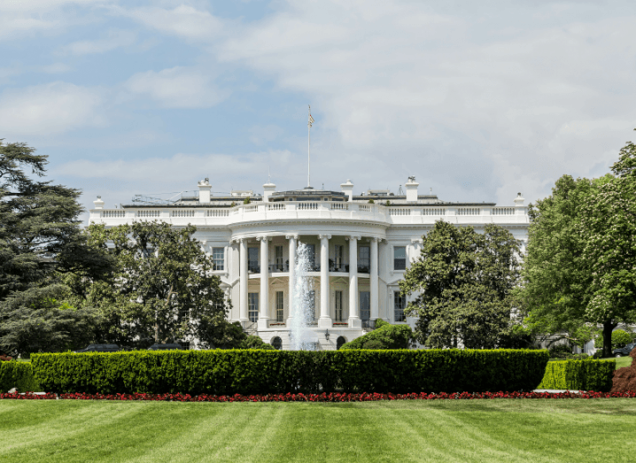 An image of the White House, a large white residential property in front of a neat lawn, with trees and hedges in front of it.