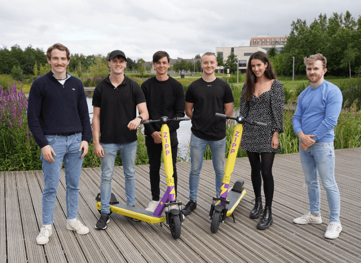 A group of young people standing on a wooden deck in front of UCD's lake. In front of the six people are two yellow e-scooters with the purple Zipp logo on them.