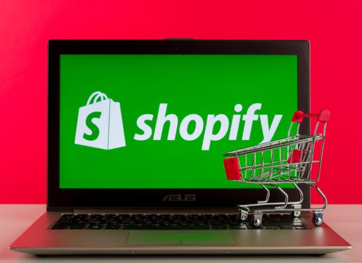 The Shopify logo is on a laptop with a toy shopping trolley against a red background.