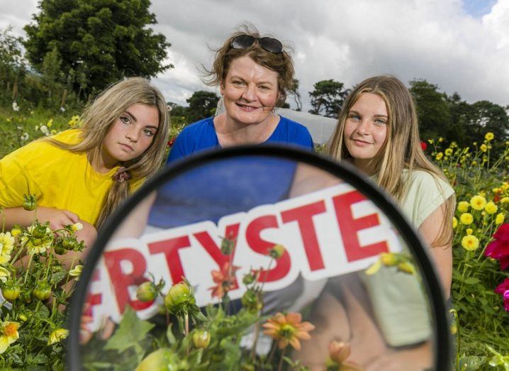 Ali Funge, Mari Cahalane and Clodagh Funge holding a BTYSTE sign in front of a microscope in a field with yellow flowers.