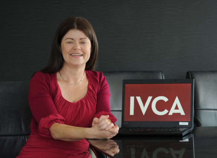 Gillian Buckley sits at a desk in a red dress, beside a laptop that says 'IVCA' on the screen.