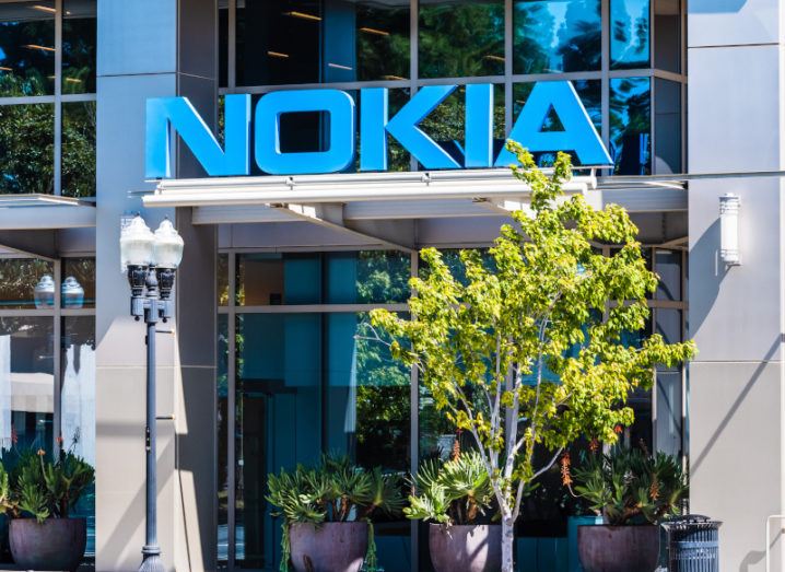 A large blue Nokia logo over the door of a glass office building on a sunny day, with a leafy tree to one side.