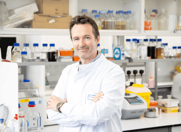 A person in a white lab coat standing with their arms crossed in a laboratory setting.