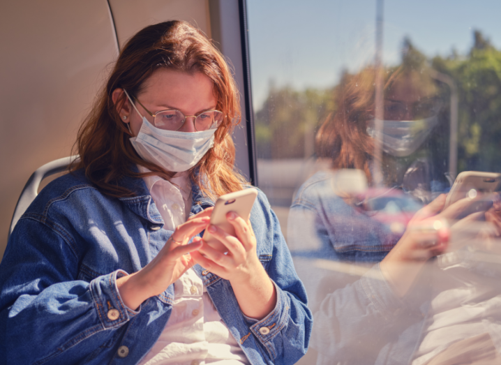 A woman wearing a denim jacket and a white mask on her face sits on a bus holding a smartphone.