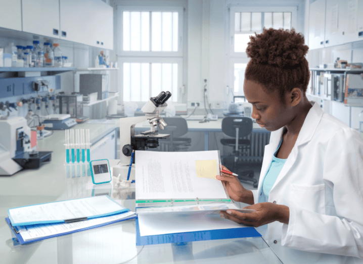 A woman in a white lab coat, sitting in a research lab reading papers.