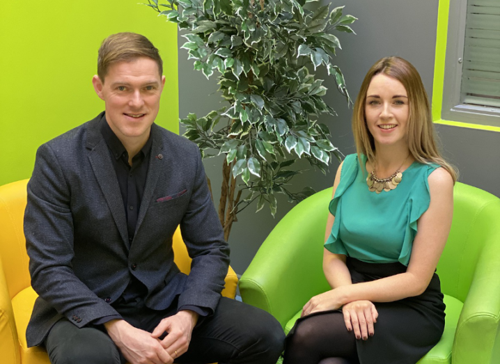 A man in a black suit sitting next to a woman wearing a green blouse and black trousers.