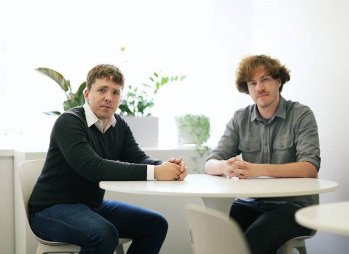 Two men in smart casual wear sit at a white table next to a window with leafy green plants on the ledge.