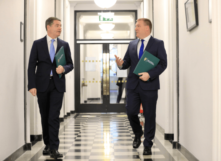 Two men walking down a corridor holding green books.