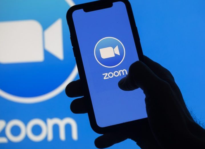 The Zoom logo on a phone in front of a larger Zoom logo on a wall.