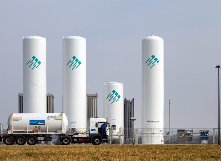 A truck transporting hydrogen fuel driving past large, white tanks filled with hydrogen fuel.