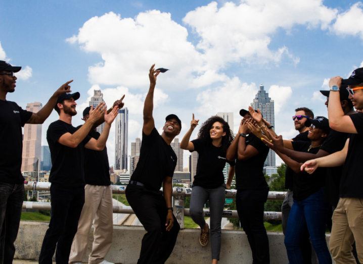 A group of people dressed in black point excitedly up to a small black square device held by a man in the centre of the picture. They are standing on a roof in front of a city and a blue sky.