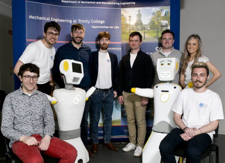 A team of seven men and one woman pose alongside two anthropomorphic white robots with smiling digital faces.