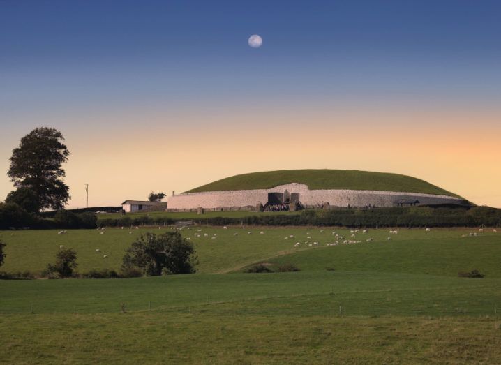 The expansive dome of the Newgrange passage tomb visible on the horizon at dusk, with the moon already visible in the darkening sky.