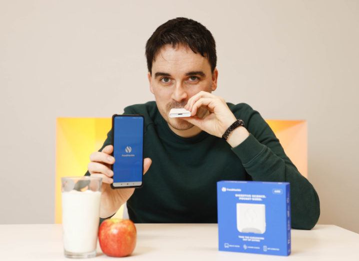 FoodMarble CEO blows into the company's Aire device, while holding a phone displaying the FoodMarble app. On the table in front of him is an apple and a glass of milk.