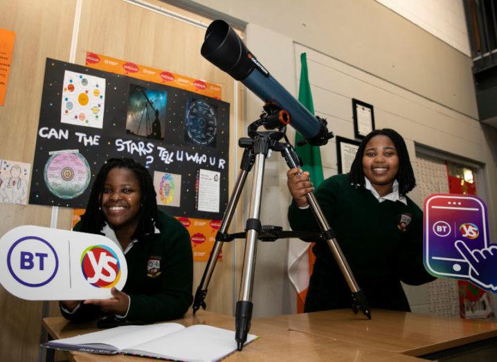 Two teenage girls in dark school uniforms stand holding a telescope and a BT Young Scientist poster. Behind them is a poster that says: ‘Can the stars tell who you are?’