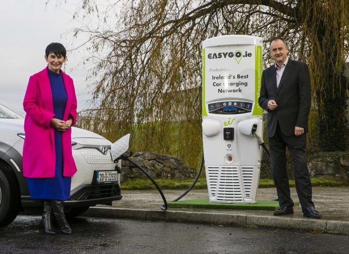 Carolan Lennon in a pink coat and Gerry Cash in a black suit standing beside an EV and charging point.