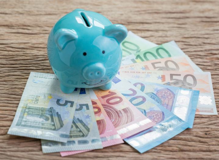 A light blue piggy bank sits on a wooden table with a variety of euro bank notes fanned out in front of it.