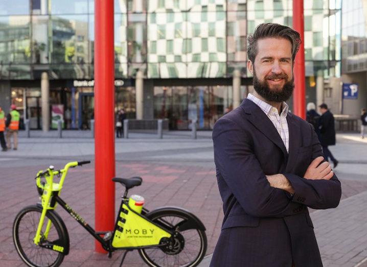 Thomas O'Connell stands in at Grand Canal Docs smiling at the camera. Behind him, a Moby-branded bike leans against a red pillar.