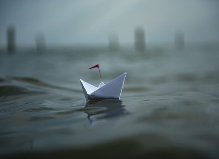 A paper boat floats on an uneasy body of water. The weather is dark and threatening.