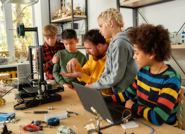 A group of children gather around an instructor in a workshop, who is showing them how to use a 3D printer.