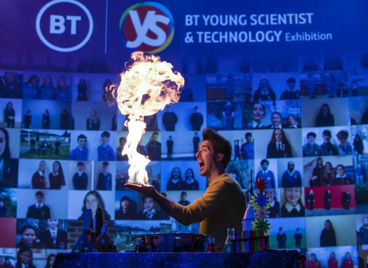 A man performing a fire trick stands in front of a large screen that says 'BT Young Scientist and Technology Exhibition' and features images of students taking part in the event.