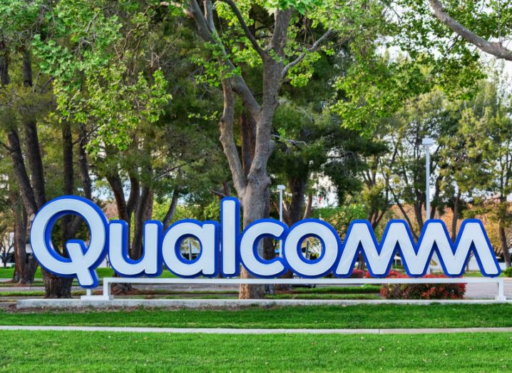 A large blue and white Qualcomm logo sign sits on the grass with trees in the background.