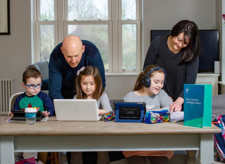 A family of five at a table. Two young girls and a boy sit at it using various devices, while their parents lean over to assist them.