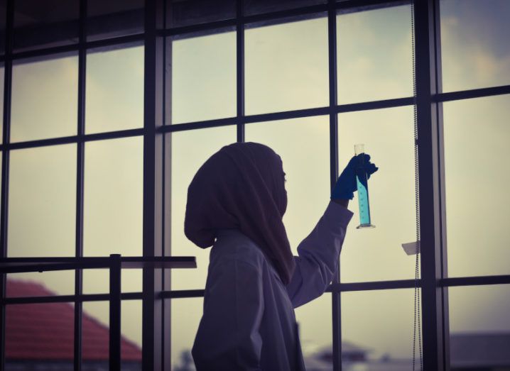 A young woman researcher is holding up a graduated cylinder in a lab with a large window.