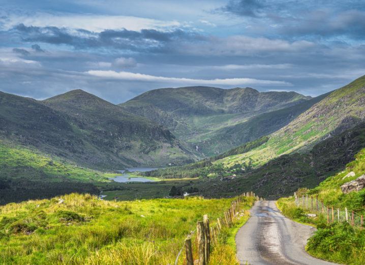 Long country road leading to The Black Valley in Co Kerry with lake and mountain range covered in sunlight and shadows from clouds.