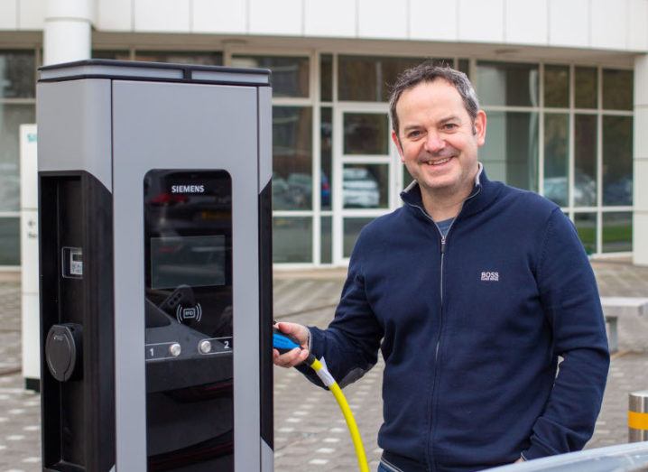 Bernard Magee stands beside a Siemens charging station for EVs, holding the plug in place.