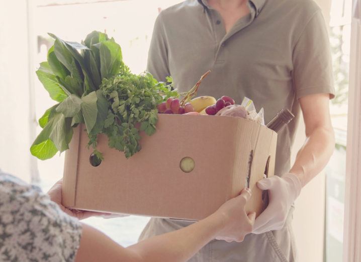 A man in a brown polo shirt is handing a cardboard box filled with fruit and vegetables to a woman.