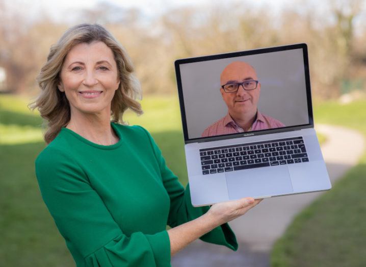 A woman in a green top stands outside holding up a laptop displaying the head and shoulders of a man wearing glasses and a pink shirt.