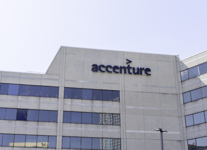 A large stone office building against a clear blue sky with the Accenture logo on the top of the building.