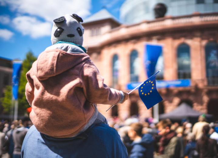 A child is held up on his father’s shoulders amid a crowd. He holds a small EU flag in his hand.