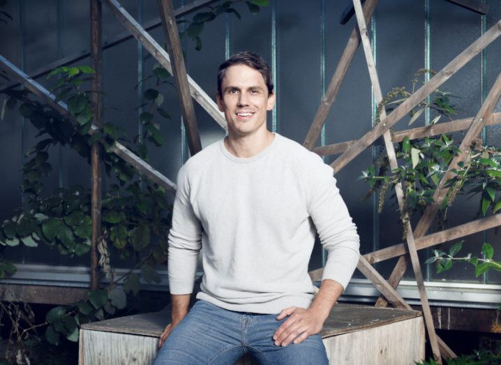 A man in jeans and a light grey sweater sits casually on a wooden platform, surrounded by green foliage.