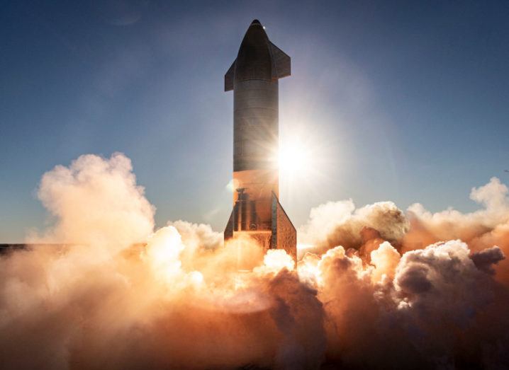 A SpaceX spacecraft takes off against a setting sun in a clear blue sky. The bottom of the rocket is engulfed in plumes of smoke.