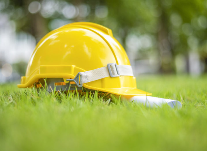 A yellow hard hat sits alongside a rolled up piece of paper on a patch of grass.