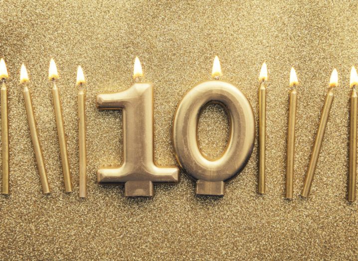 A row of 10 lit golden candles on a glittering background. The candles in the centre are shaped like the number 10.