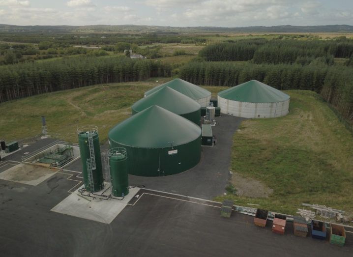 An aerial view of a large green biogas production facility, comprising several green round buildings on a large site.