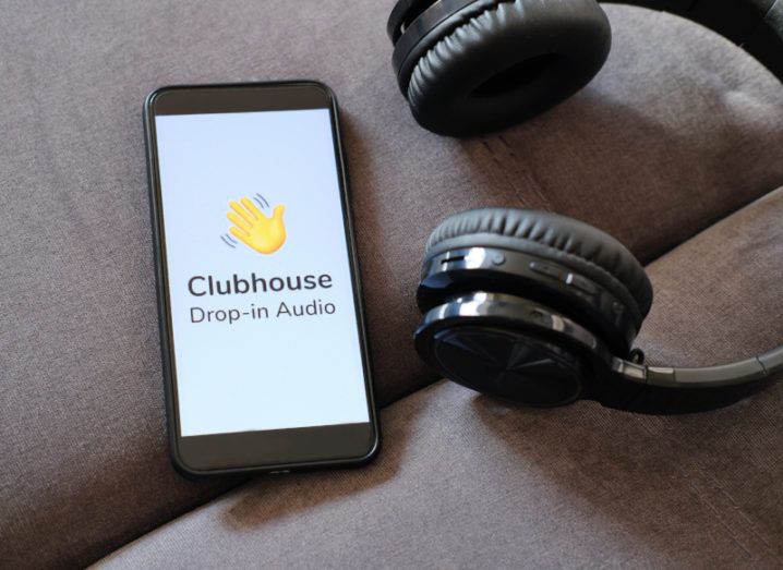 A smartphone sits on a brown couch next to a pair of black headphones. The Clubhouse logo is on the phone’s screen.