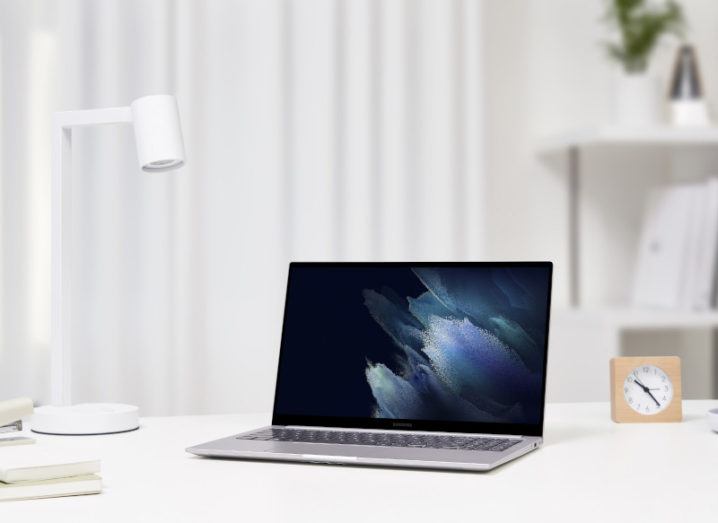 A new Samsung Galaxy Book laptop is sitting on a white desk, beside a lamp and a clock.