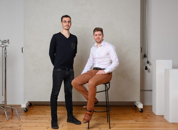 Two men are in a studio. One is standing and one is sitting on a stool.