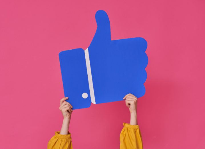 Hands are holding up a large cutout of a thumbs-up emoji, resembling the Facebook ‘like’ icon.