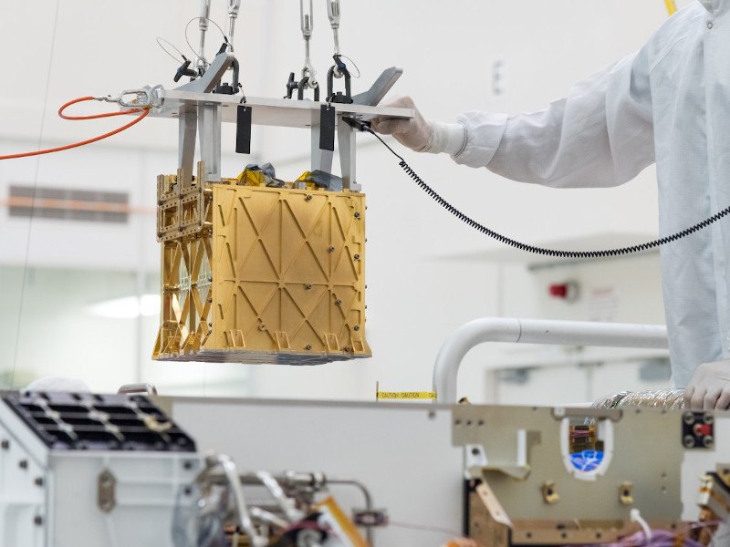 A small gold box is being lowered into a NASA rover in a lab setting.