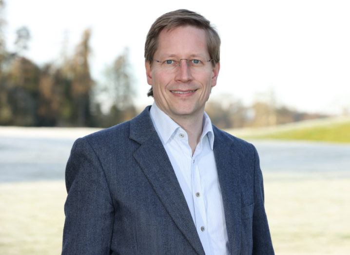 Holger Claussen wears a blue-grey twill blazer and white shirt. He is pictured outdoors, with a blurred green landscape in the background.