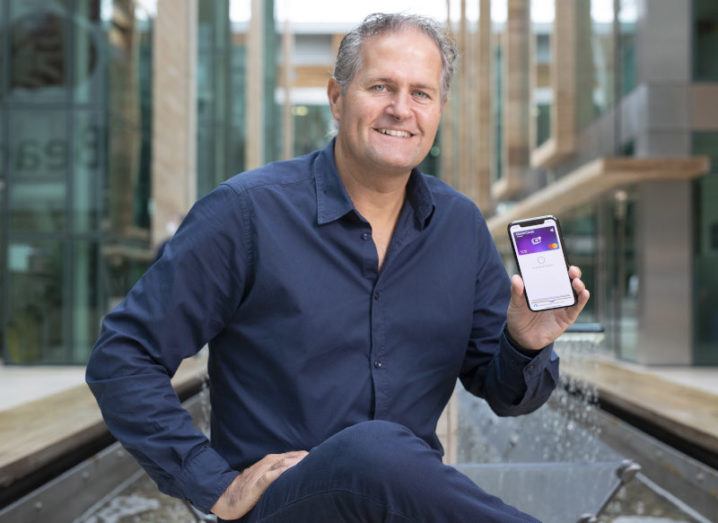 CleverCards founder and CEO Kealan Lennon is sitting outdoors in a modern business district and smiling into the camera. He is holding up a phone with the CleverCards app open.