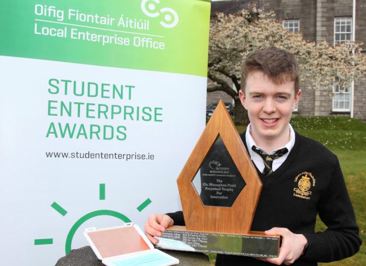 Student Matthew McVicar holds a large wooden award, while standing beside his CopperCase invention in front of a sign that reads 'Student Enterprise Awards'.